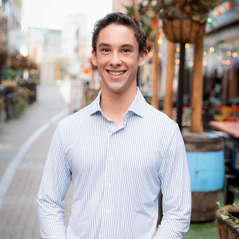 A young man with dark hair, wearing a blue and white striped button-down shirt, smiles warmly at the camera. He is standing outdoors on a city street.