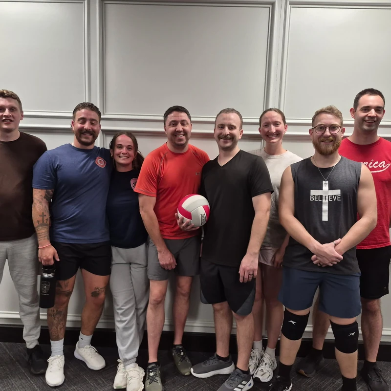 A group of eight smiling people in athletic wear pose together for a photo. A man in the center is holding a red and white volleyball.