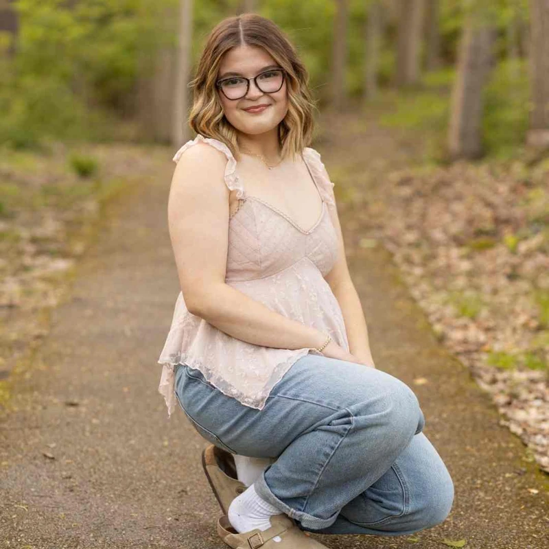 A young woman with glasses and wavy brown hair smiles while crouching on a paved path in a wooded area. She is wearing a pink lace top and blue jeans.