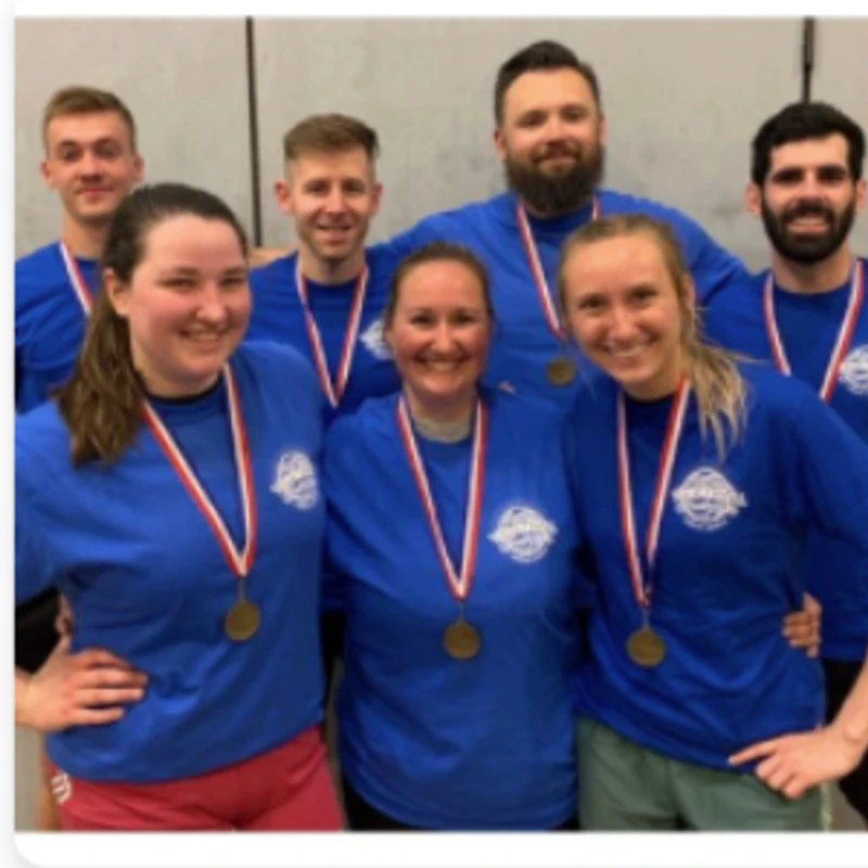 A group of seven smiling adults on a sports team, wearing matching blue shirts and medals, posing for a celebratory group photo after a victory.