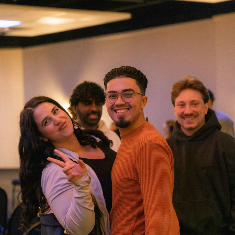 A group of four young, diverse friends smile for the camera at an event. A woman on the left makes a peace sign while a man in an orange sweater smiles widely.