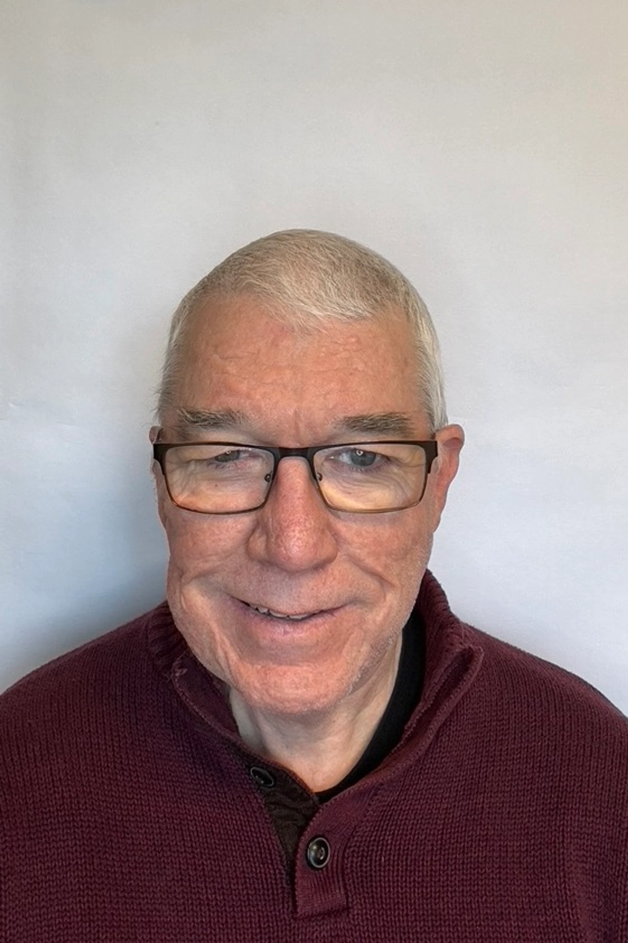 A headshot of an older man with short gray hair and glasses. He is smiling and wearing a maroon knit sweater against a plain white background.