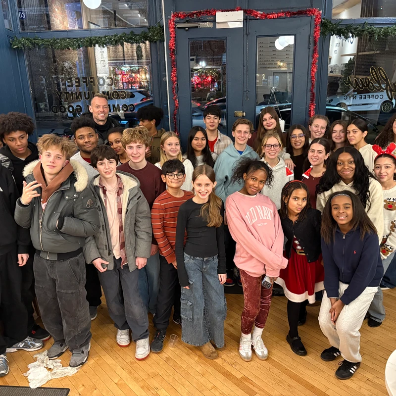 A large, diverse group of teenagers and a few adults pose together for a photo in a cafe decorated for the holidays with tinsel and garland.