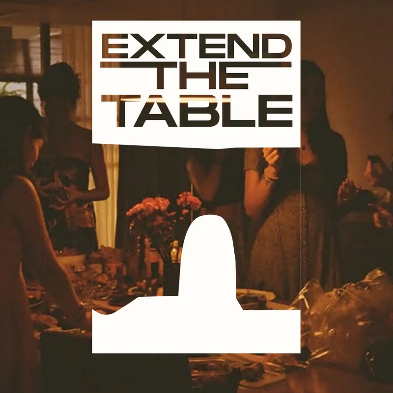 A group of people gathers around a dinner table in a warmly lit room. A graphic overlay reads 'Extend The Table' in bold, stylized letters.