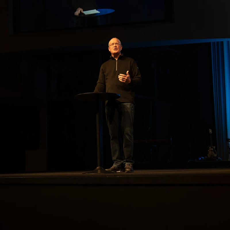 An older man with glasses stands on a dark stage speaking at a lectern. A large screen above him shows a close-up of him also speaking.