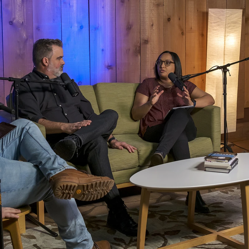 Three people sit in a cozy, wood-paneled room recording a podcast. A man and a woman on a couch speak into microphones while another person listens.