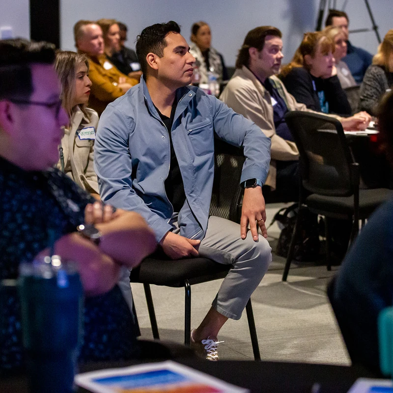 A diverse group of people sit in an audience at a professional conference, looking attentively towards a speaker or screen just out of frame.