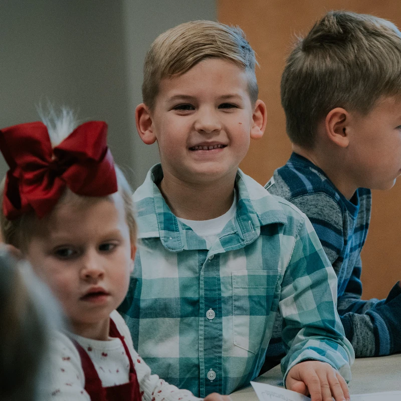 A young boy with blond hair in a teal plaid shirt smiles at the camera. He is sitting at a table with other children, one a girl with a large red bow.