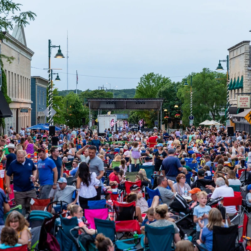 A large crowd of people gathered on a downtown street for an outdoor concert. People sit in lawn chairs watching a band perform on a stage in the distance.