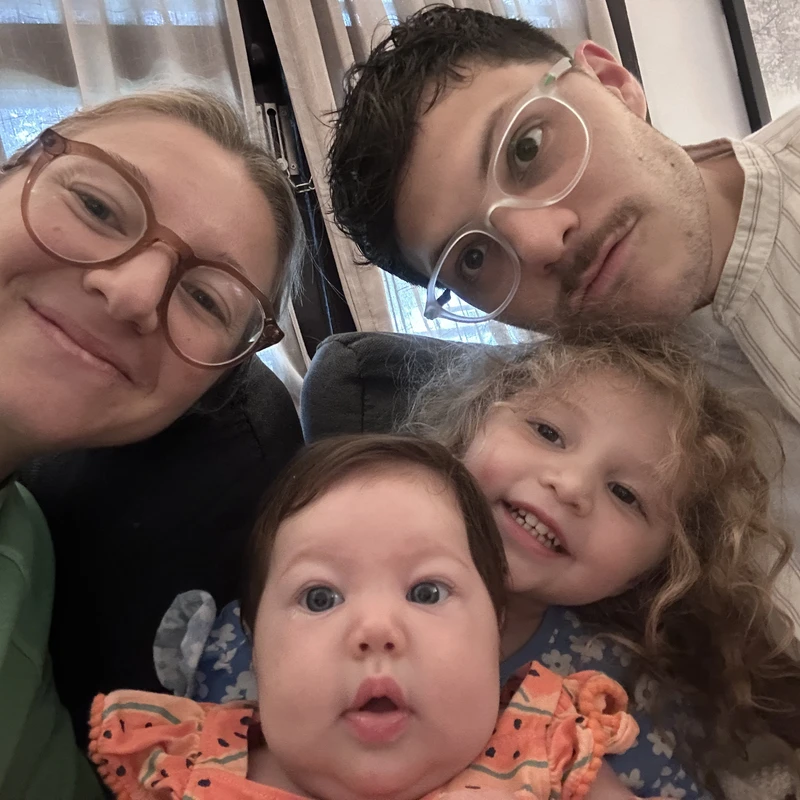 A close-up selfie of a smiling family of four. A mother and father with glasses pose with their two young daughters, a toddler and a baby.