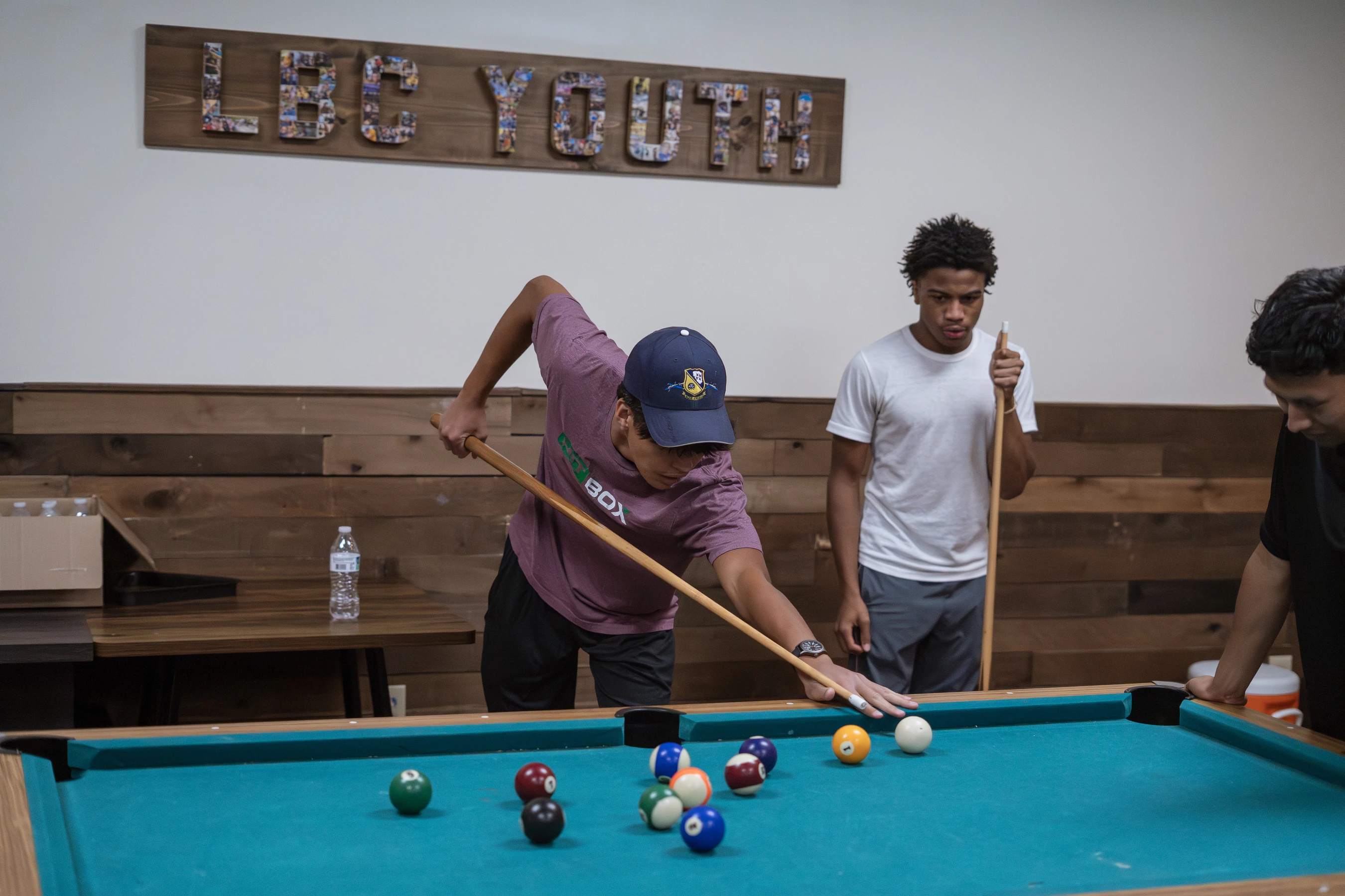 Three young men gather around a teal pool table. A man in a cap and purple shirt leans in to take his shot as his friends watch.