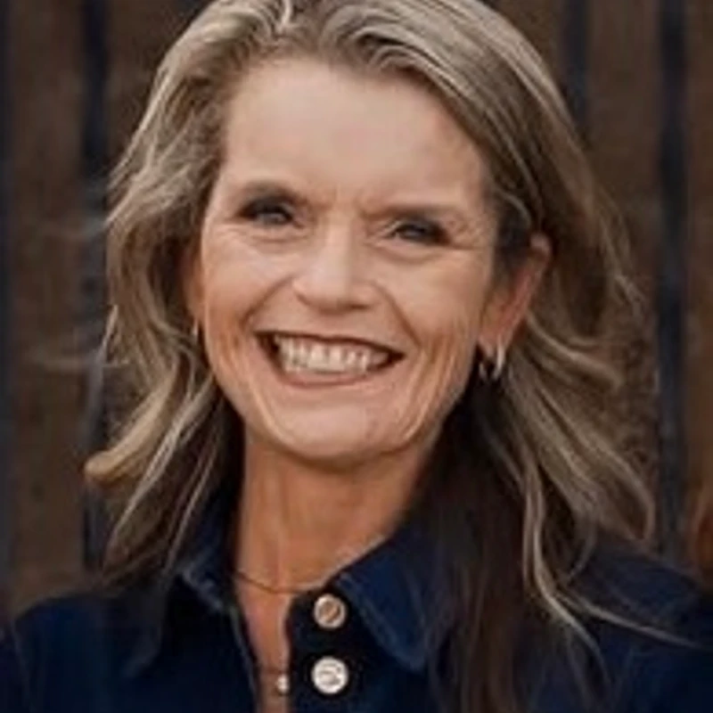 A friendly, smiling woman with blonde, shoulder-length hair poses for a headshot wearing a dark blue collared shirt.