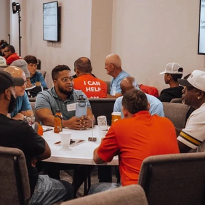 A diverse group of men of various ages sit at round tables in a conference room, engaged in a discussion during a church meeting or event.