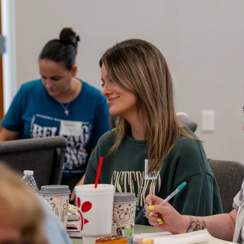 A young woman in a green sweatshirt smiles while sitting at a table with other attendees during a meeting, with notebooks and drinks in front of them.