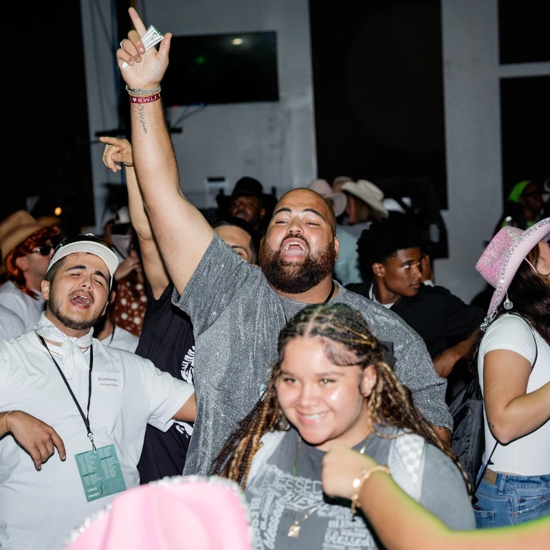 A diverse crowd of young people dance and sing at a party. A man in a sparkly shirt is in the center, cheering with his arm raised high among his friends.
