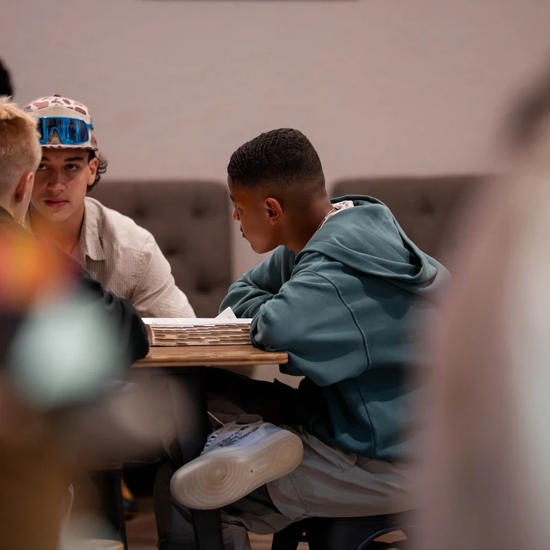 Three young men sit at a wooden table, engaged in a serious conversation. One wears a hoodie, another a cap with sunglasses, and a third has his back to us.