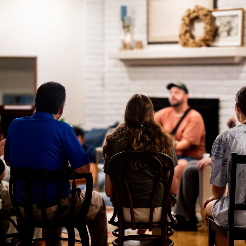 A group of people sitting on chairs with their backs to the camera, listening to a speaker in front of a white brick fireplace in a cozy home.