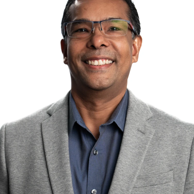 Headshot of a smiling man with dark hair and glasses, wearing a blue collared shirt and a grey blazer against a white background.