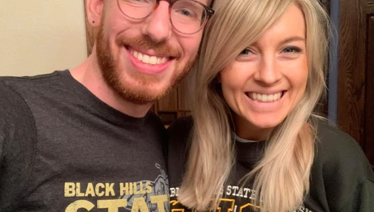 A man with red hair and glasses and a woman with blonde hair smile for a photo while both wearing Black Hills State University shirts.