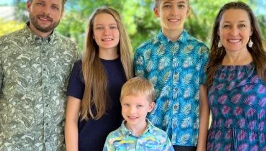 A smiling family of five, with two parents and three children, stands together for a portrait outdoors in front of green trees.