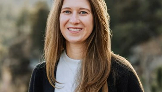 A headshot of a smiling woman with long, light brown hair. She is wearing a white shirt and a black cardigan and is standing outdoors.