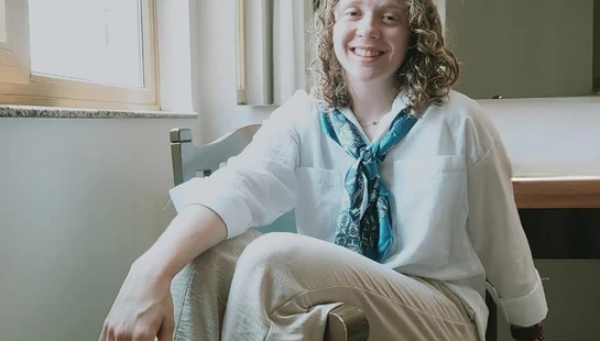 A person with curly blonde hair smiles while sitting on a green chair. They wear a white shirt, beige pants, and a blue paisley scarf around their neck.