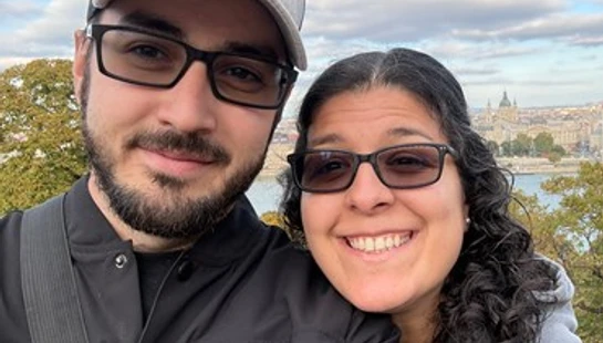A smiling man and woman take a selfie with a scenic city and river view behind them. The man wears a hat and glasses, and the woman rests on his shoulder.