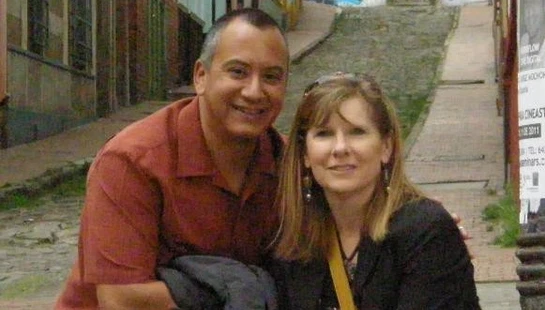 A smiling man and woman pose together on a narrow, cobblestone city street lined with colorful buildings. Graffiti is visible on a brick wall in the background.