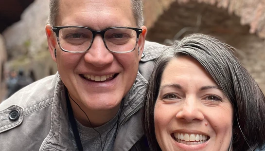 A close-up selfie of a smiling man and woman. He wears glasses and a grey jacket, and she has dark hair with grey streaks. Both are wearing earbuds.