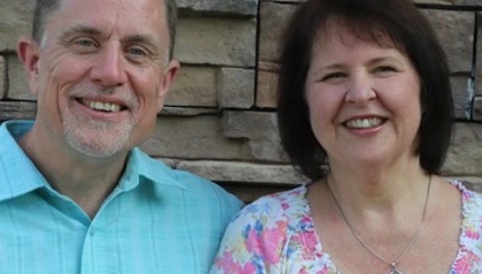 A man in a light blue shirt and a woman in a floral blouse stand side-by-side, smiling for a portrait against a stacked stone wall background.