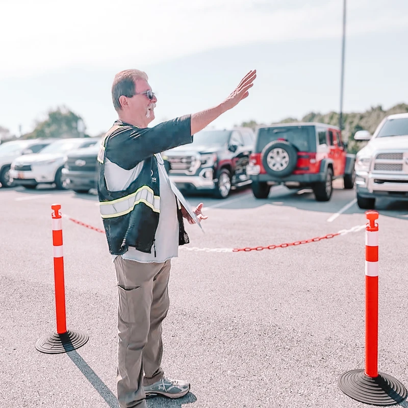 A man in a safety vest and sunglasses smiles while directing cars with a wave of his arm in a full parking lot on a sunny day.