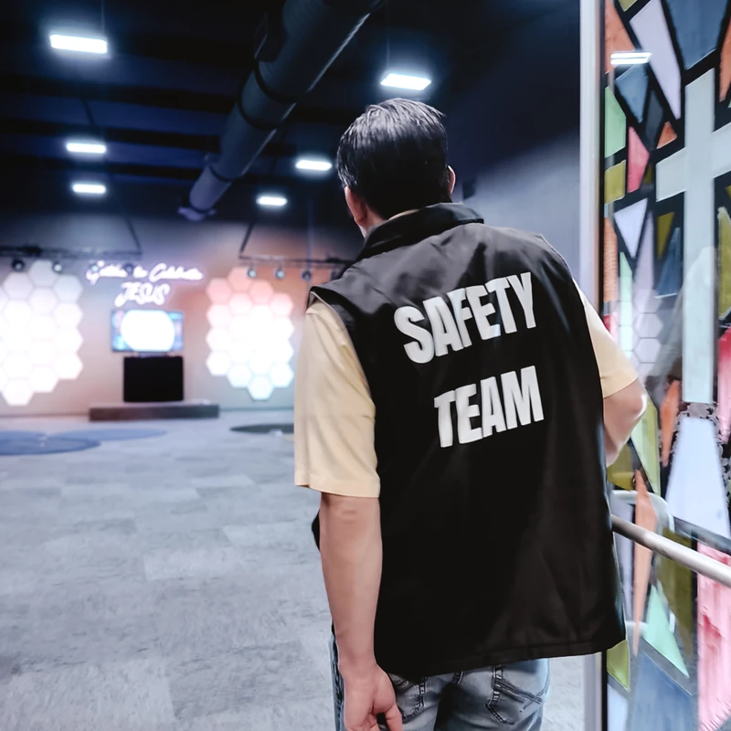 A man from behind wearing a black 'Safety Team' vest looks into a large, modern church sanctuary with a stage and lit geometric panels in the background.