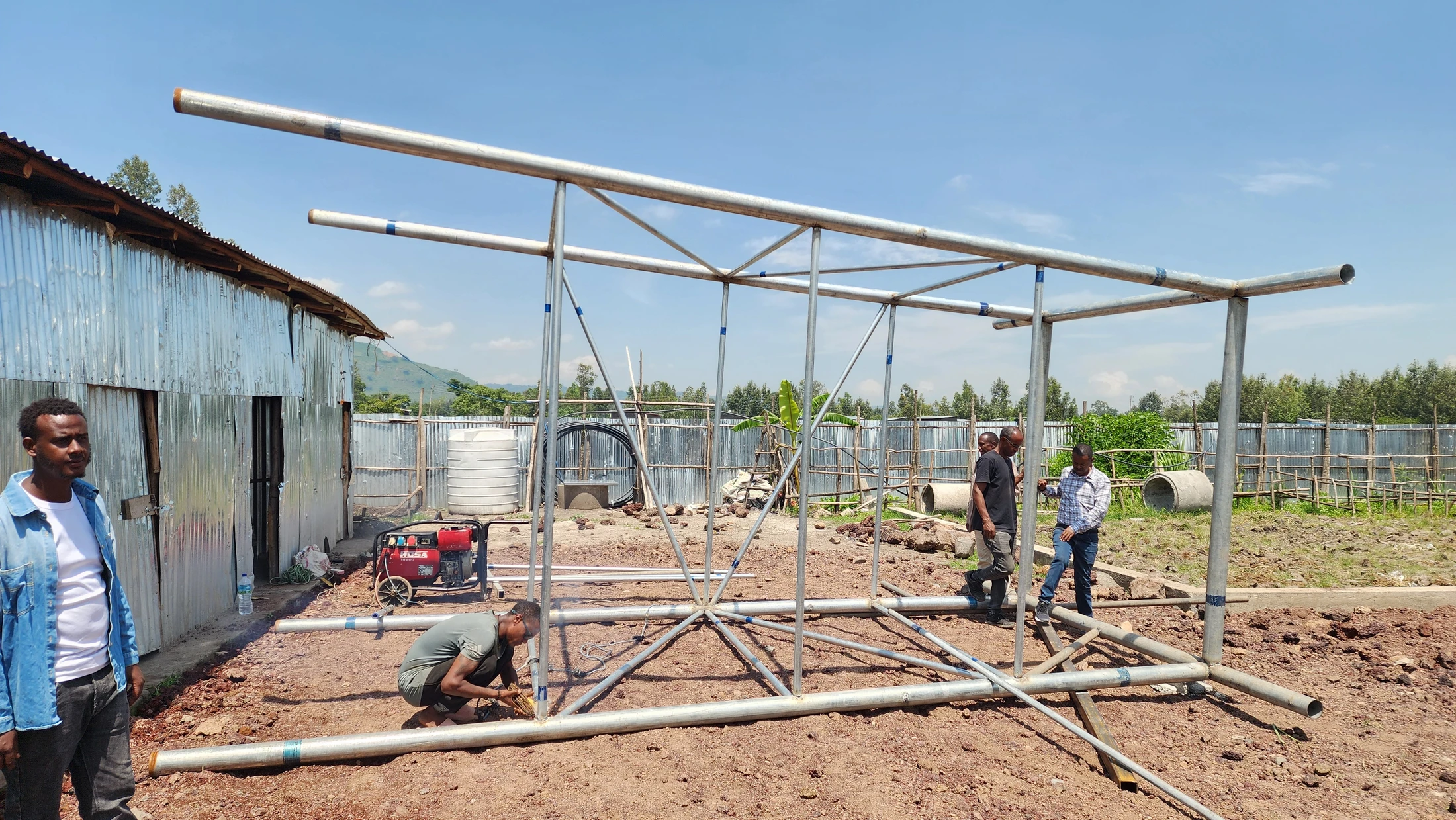A group of men assemble a large, tubular metal frame on a dirt lot. One man kneels to weld a joint while others look on under a sunny, blue sky.