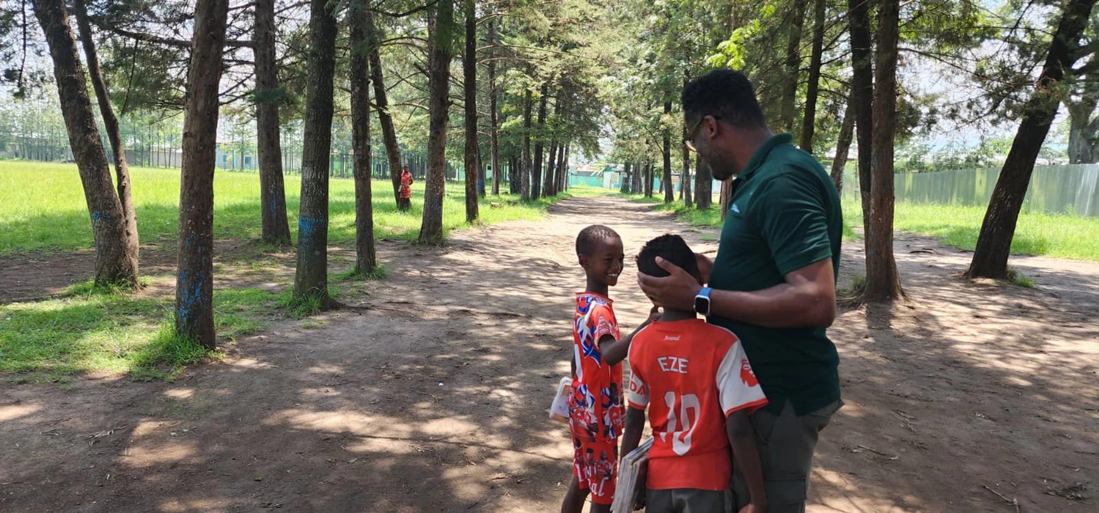 A man in a green shirt gently holds the head of a young boy while another boy stands beside them smiling. They are on a dirt path lined with tall trees.