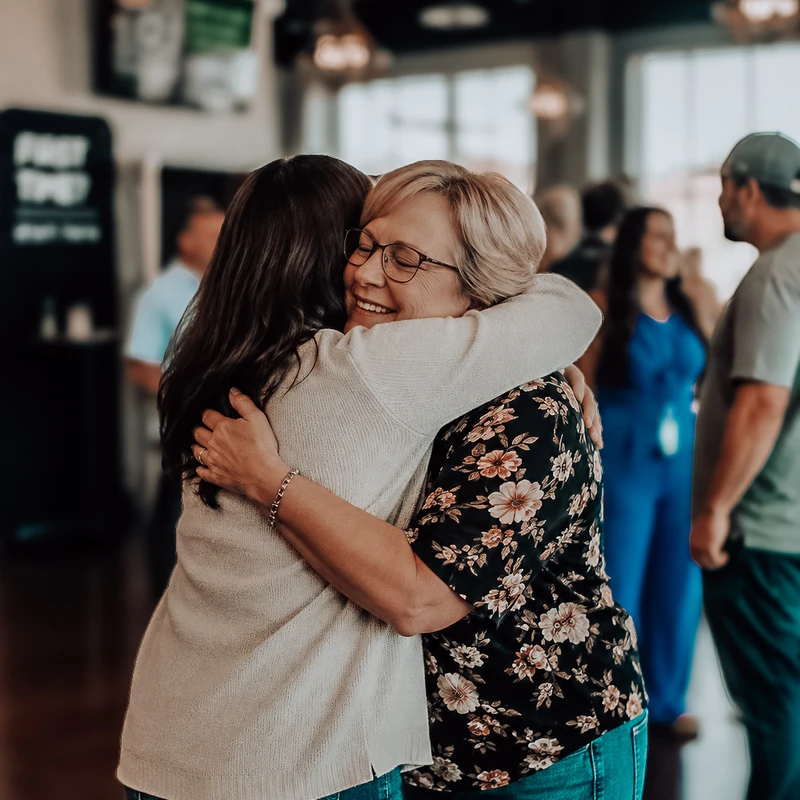 Two women share a warm hug in a room full of people. The older woman, with blonde hair and glasses, smiles with her eyes closed as she embraces the other woman.