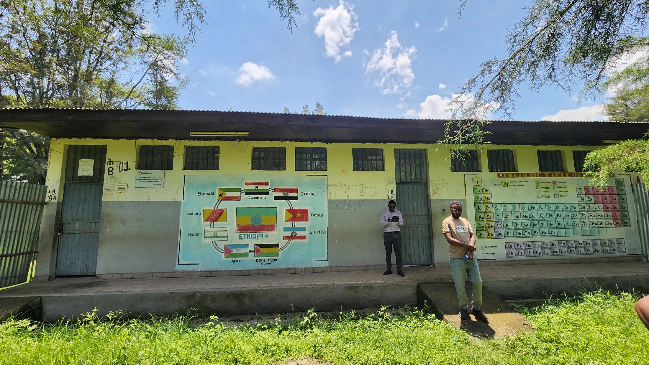 Two men stand outside a rural school building in Ethiopia, which has murals of the periodic table and a map of Ethiopia painted on its exterior wall.