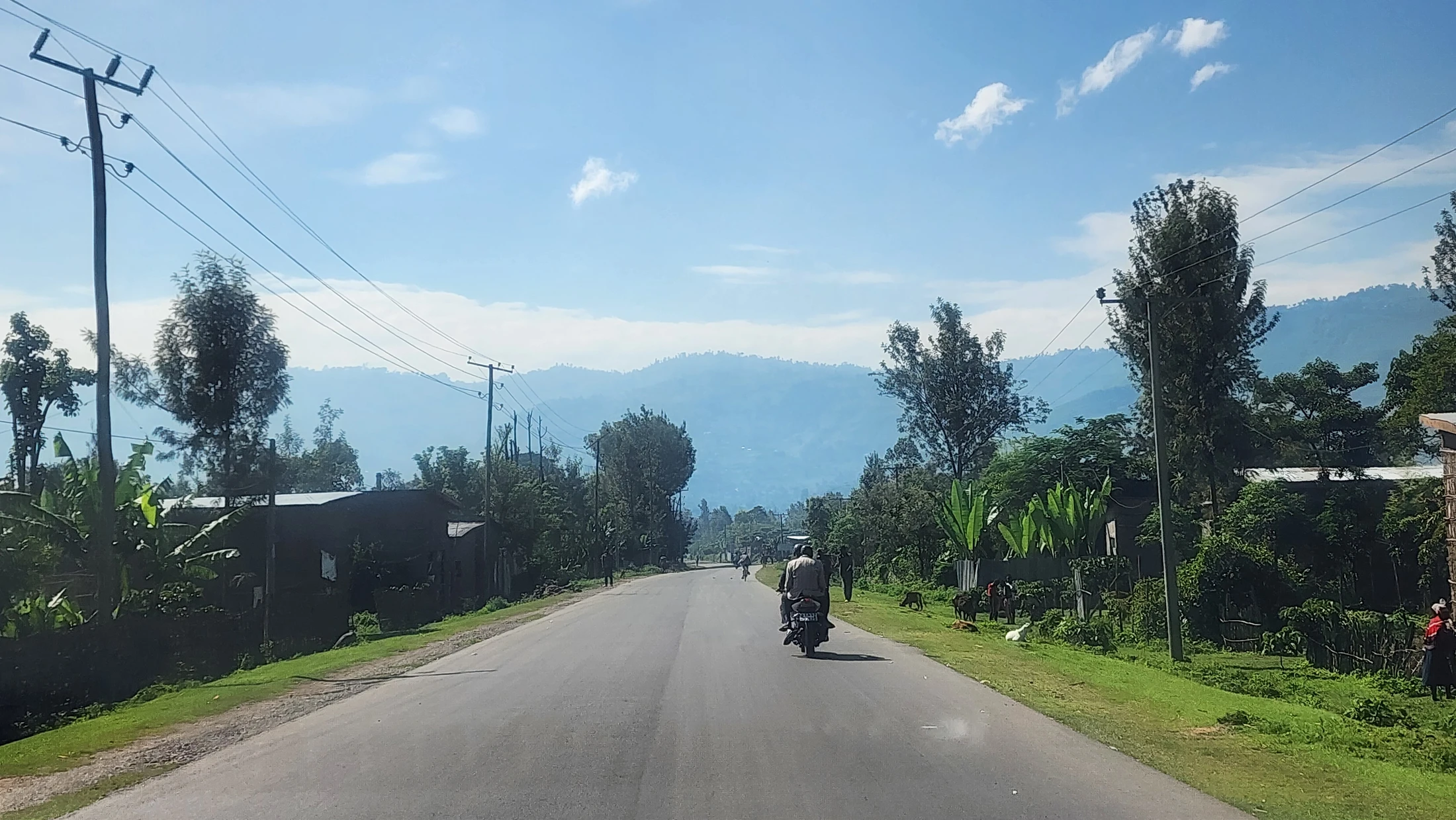 A person rides a motorcycle down a paved road through a lush, green rural village with hazy blue mountains visible in the distance under a clear sky.