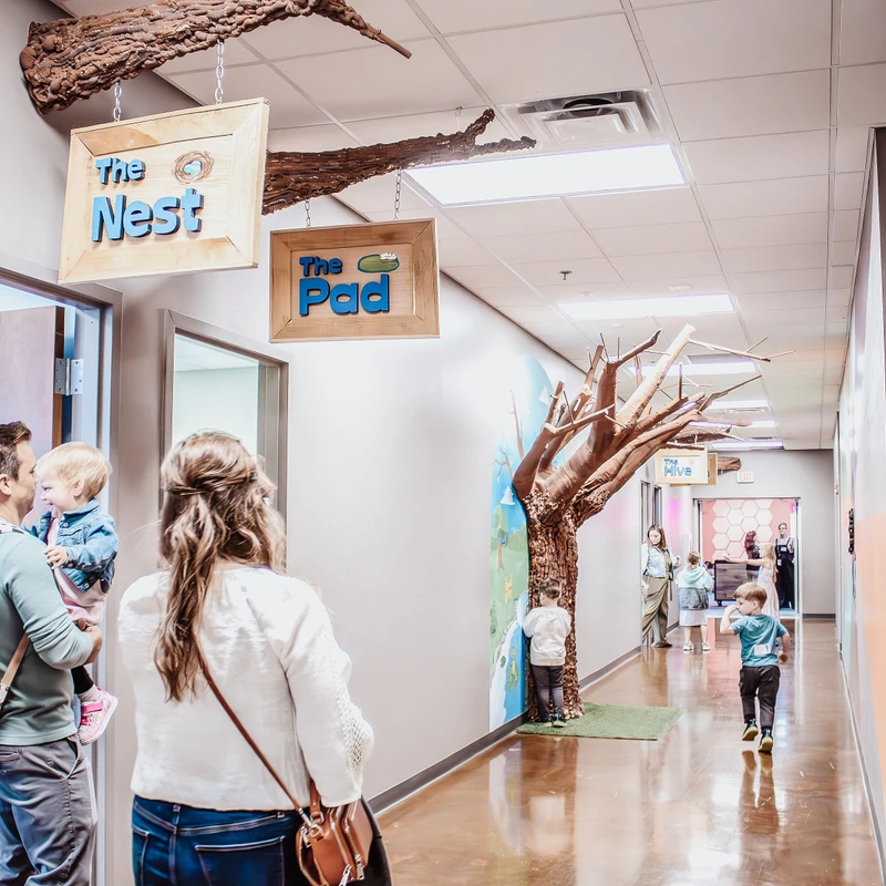 A family stands in a brightly lit hallway of a children's center. The hall has nature-themed decor, including signs for 'The Nest' and 'The Pad'.