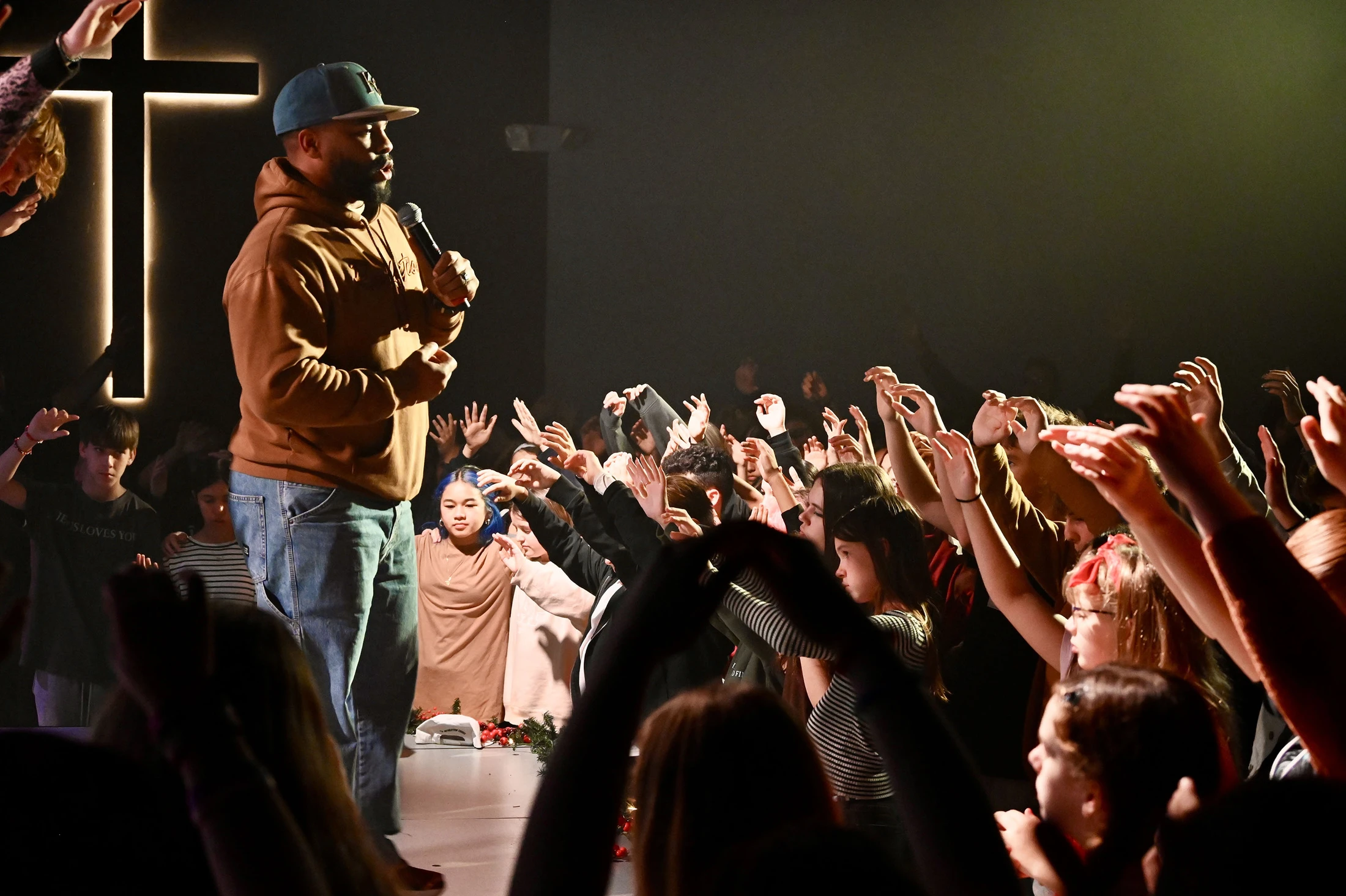 A man in a hoodie and cap speaks on stage to a crowd of young people with their hands raised in worship. A large, lit cross is in the background.