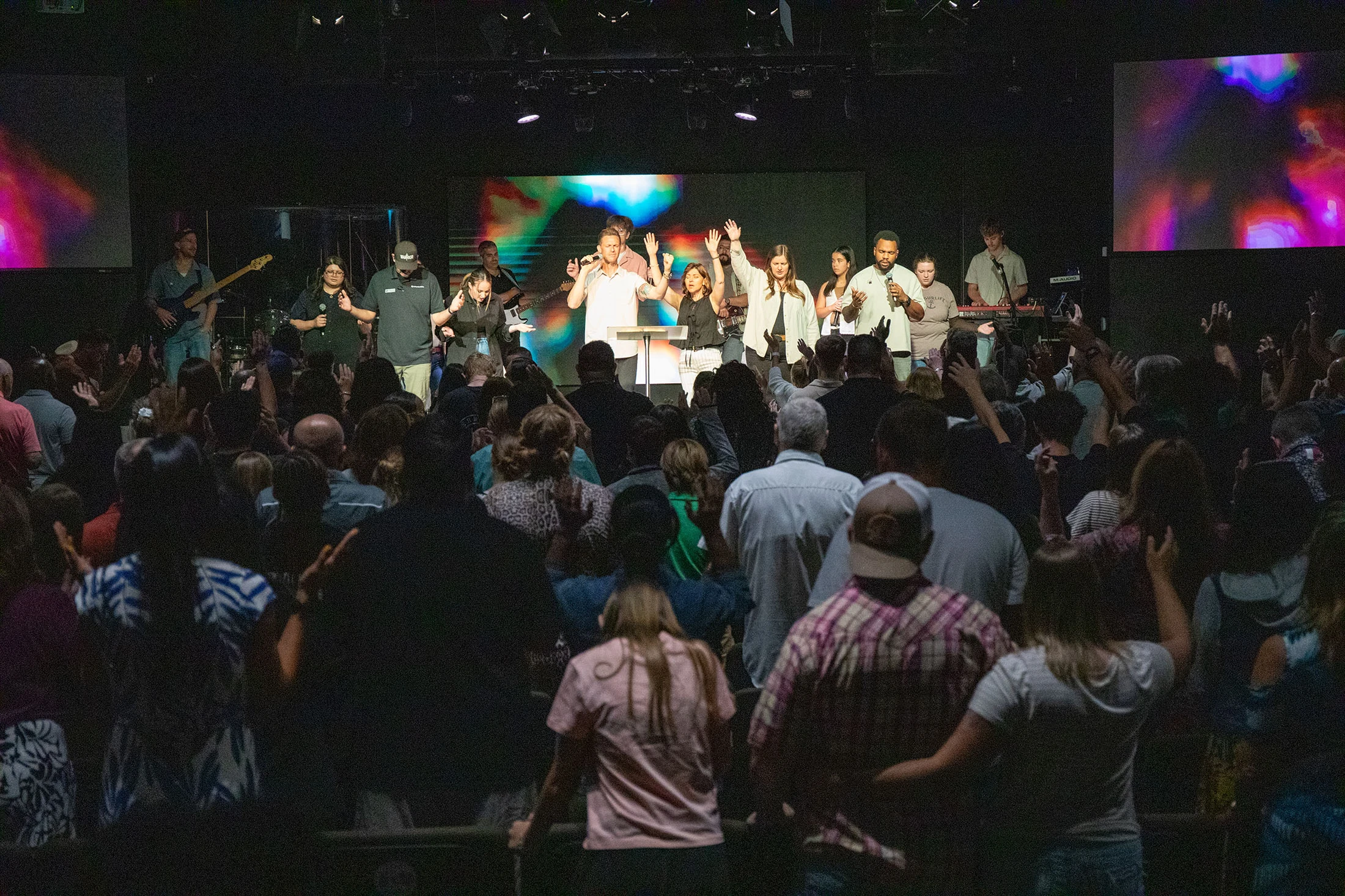 A view from the back of a large crowd raising their hands in worship toward a diverse band and singers performing on a brightly lit stage in an auditorium.