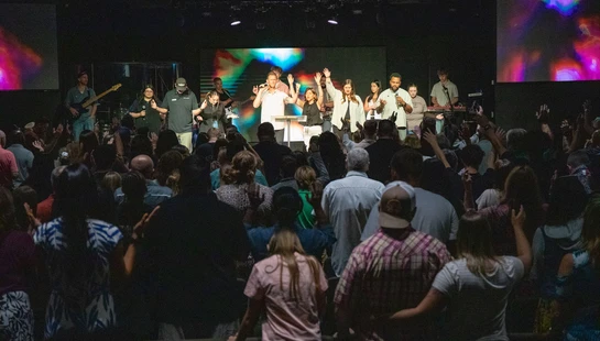 A view from the back of a large crowd raising their hands in worship toward a diverse band and singers performing on a brightly lit stage in an auditorium.
