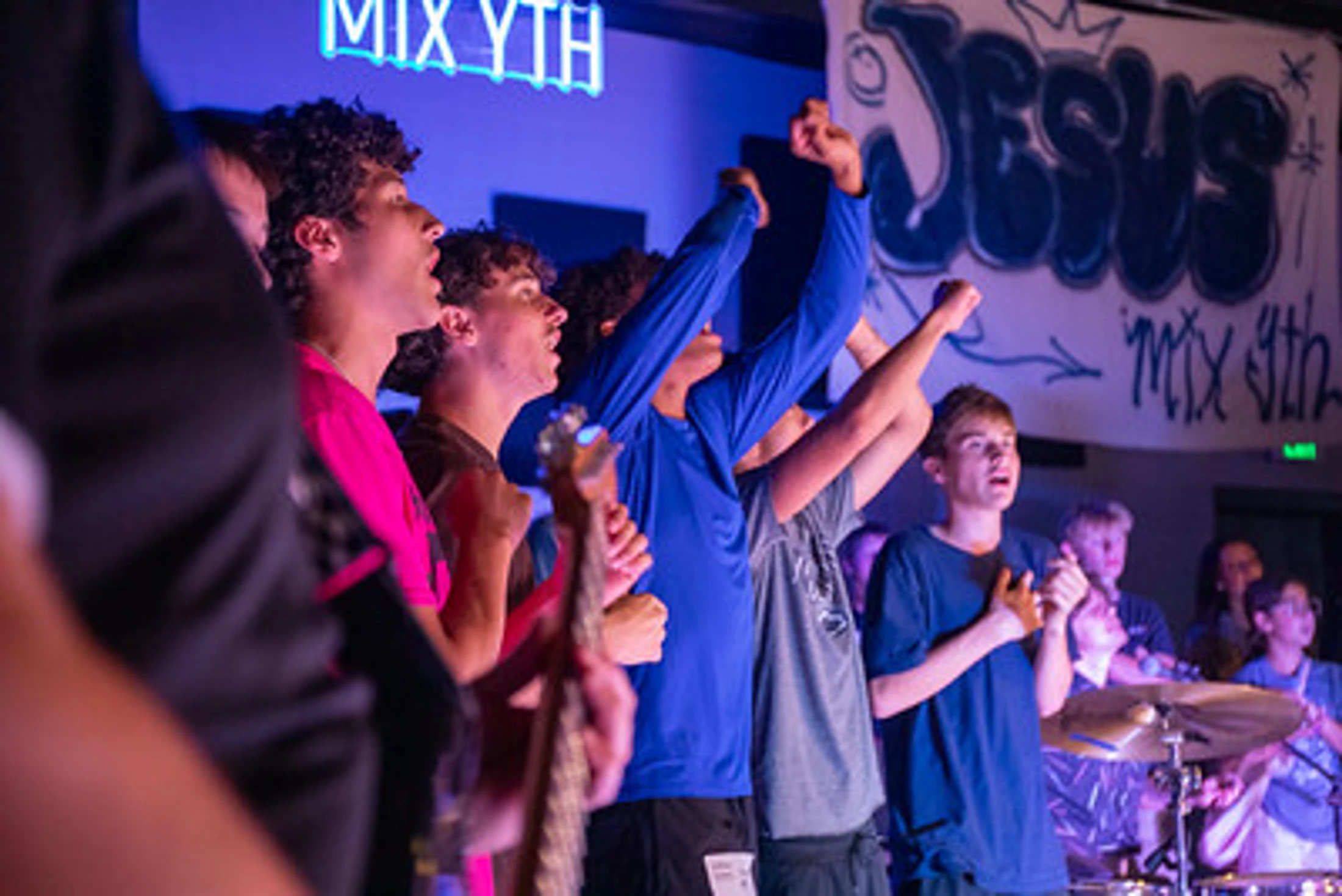 A group of teenage boys stand with hands raised, singing enthusiastically under blue lights. A banner with 'JESUS' in graffiti letters hangs behind them.