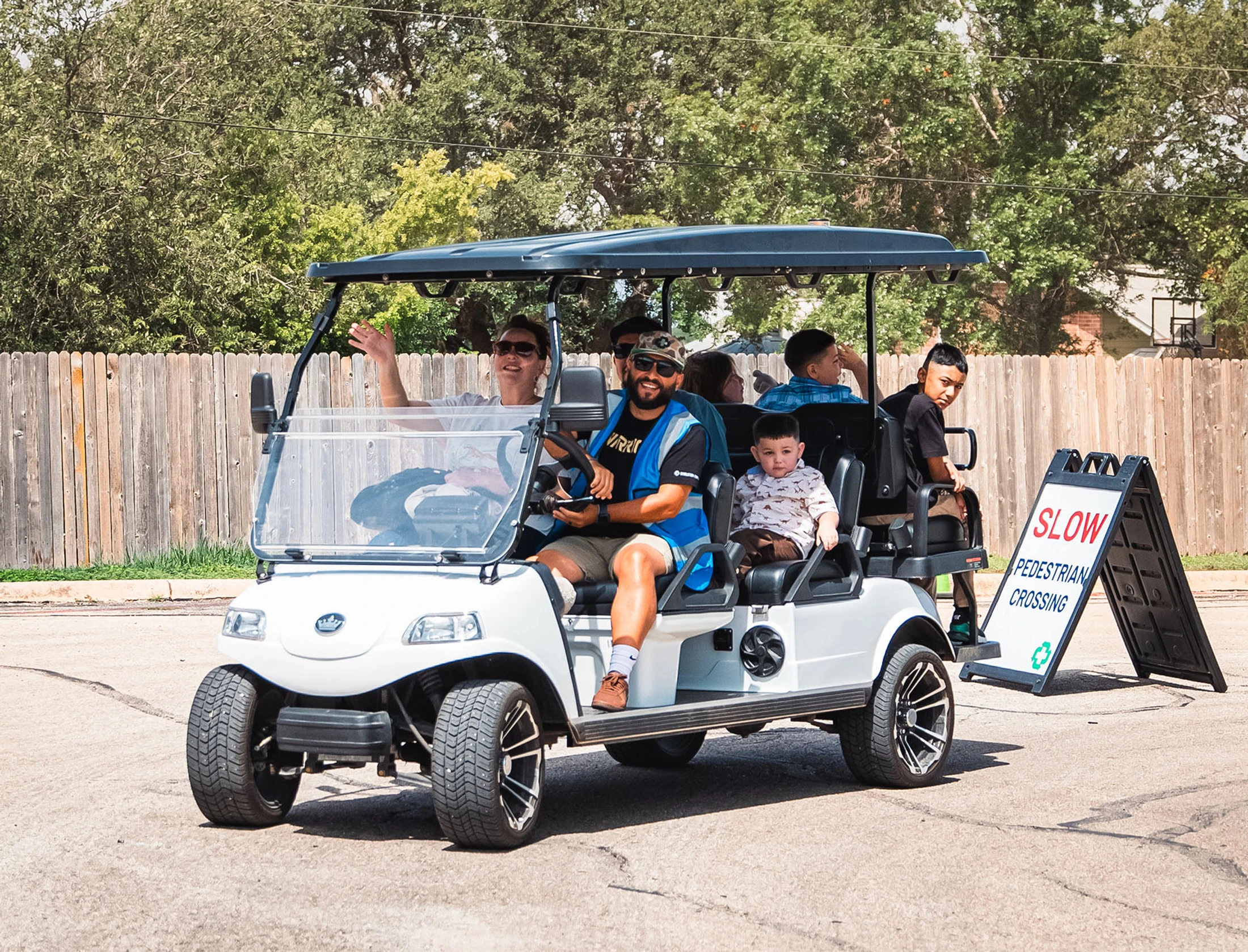 A smiling man in a blue vest drives a white golf cart with several adults and children as passengers on a paved road next to a pedestrian crossing sign.