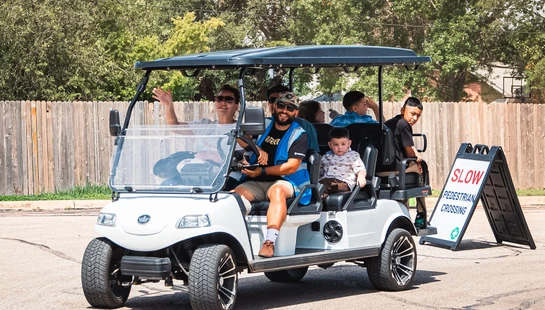 A smiling man in a blue vest drives a white golf cart with several adults and children as passengers on a paved road next to a pedestrian crossing sign.