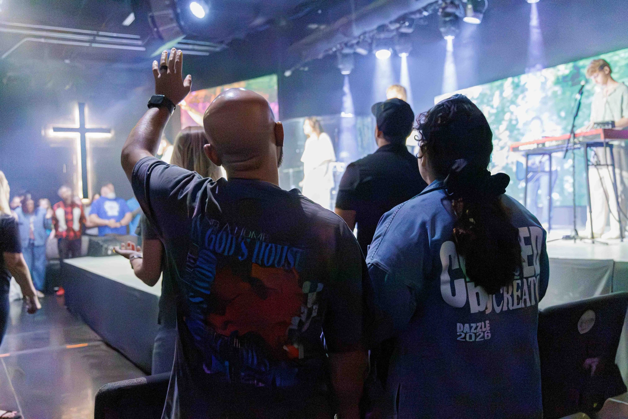 A bald man, seen from behind, raises his hand in worship during a church service. A band is on stage under bright lights, and a glowing cross is visible.