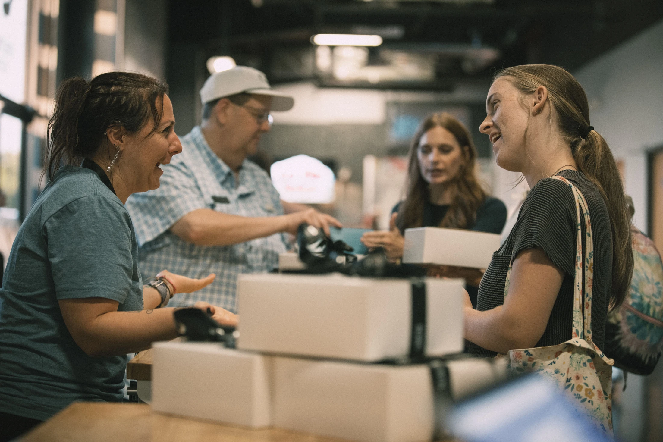 A female volunteer smiles as she talks to a young woman across a counter with white boxes. Other people are in the background at an indoor event.
