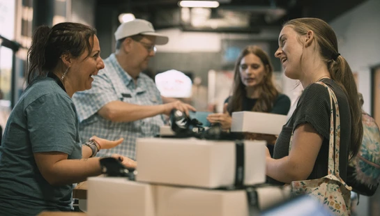 A female volunteer smiles as she talks to a young woman across a counter with white boxes. Other people are in the background at an indoor event.
