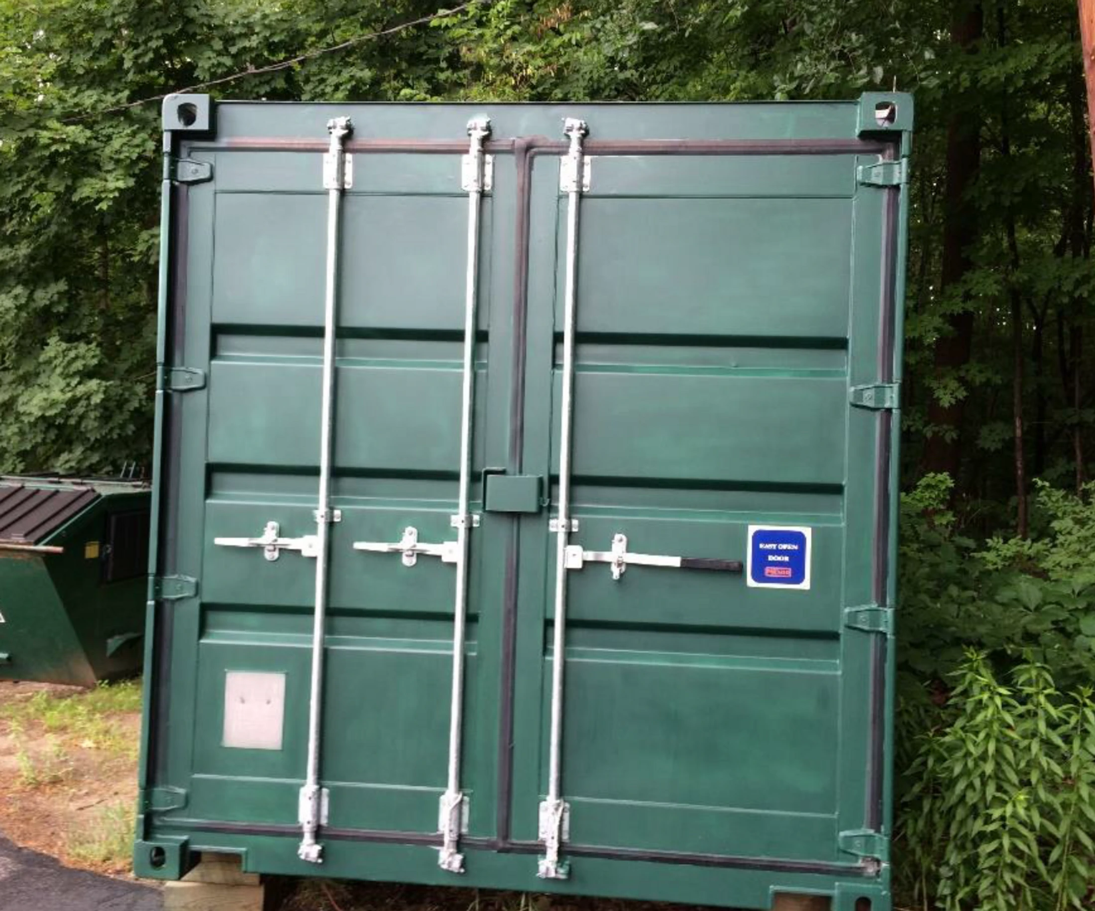 The locked double doors of a dark green shipping container standing on wooden blocks in front of a line of green trees. A green dumpster is visible to the left.