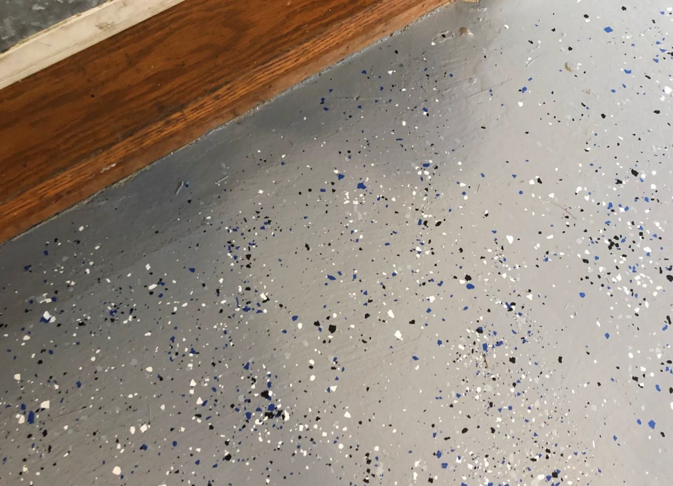 A close-up view of a gray epoxy floor speckled with blue, black, and white decorative flakes, with a wooden door threshold visible at the top of the frame.