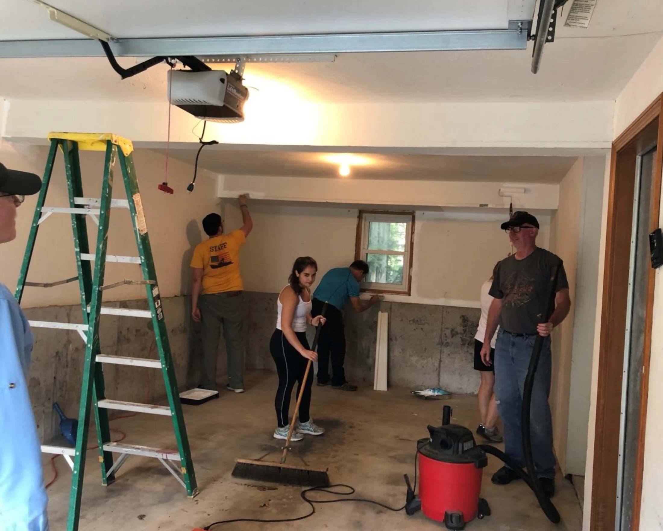 A group of people work together in a garage, painting walls and trim, sweeping the floor, and using a large shop-vac to clean during a renovation project.